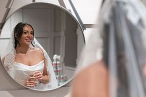 Bride in white wedding dress and veil admires her reflection in a round mirror while getting ready.