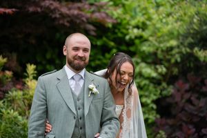 Laughing bride in lace veil and groom in sage green kilt suit pose together outdoors at a garden wedding.