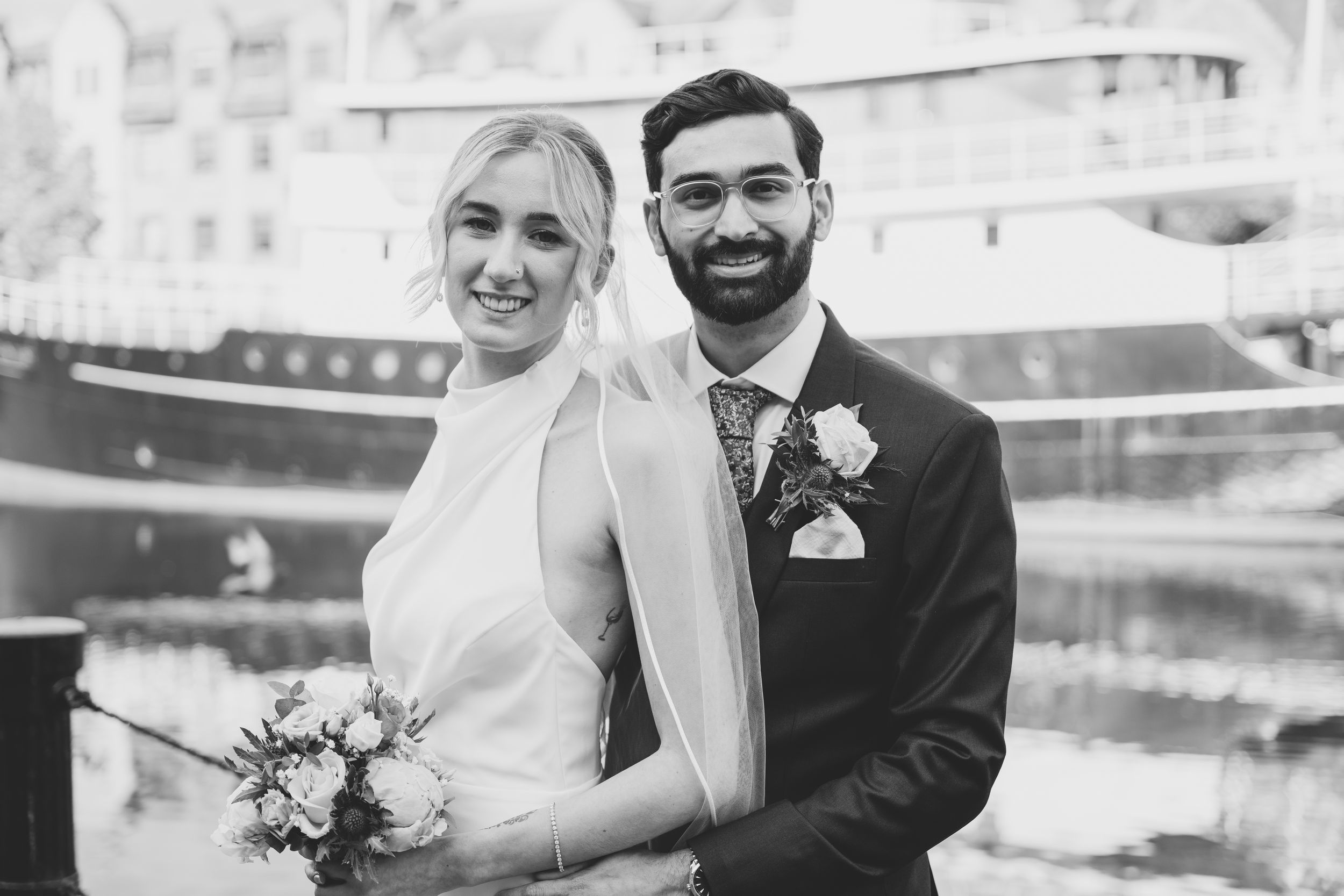 Wedding couple posing together near waterfront with urban backdrop.