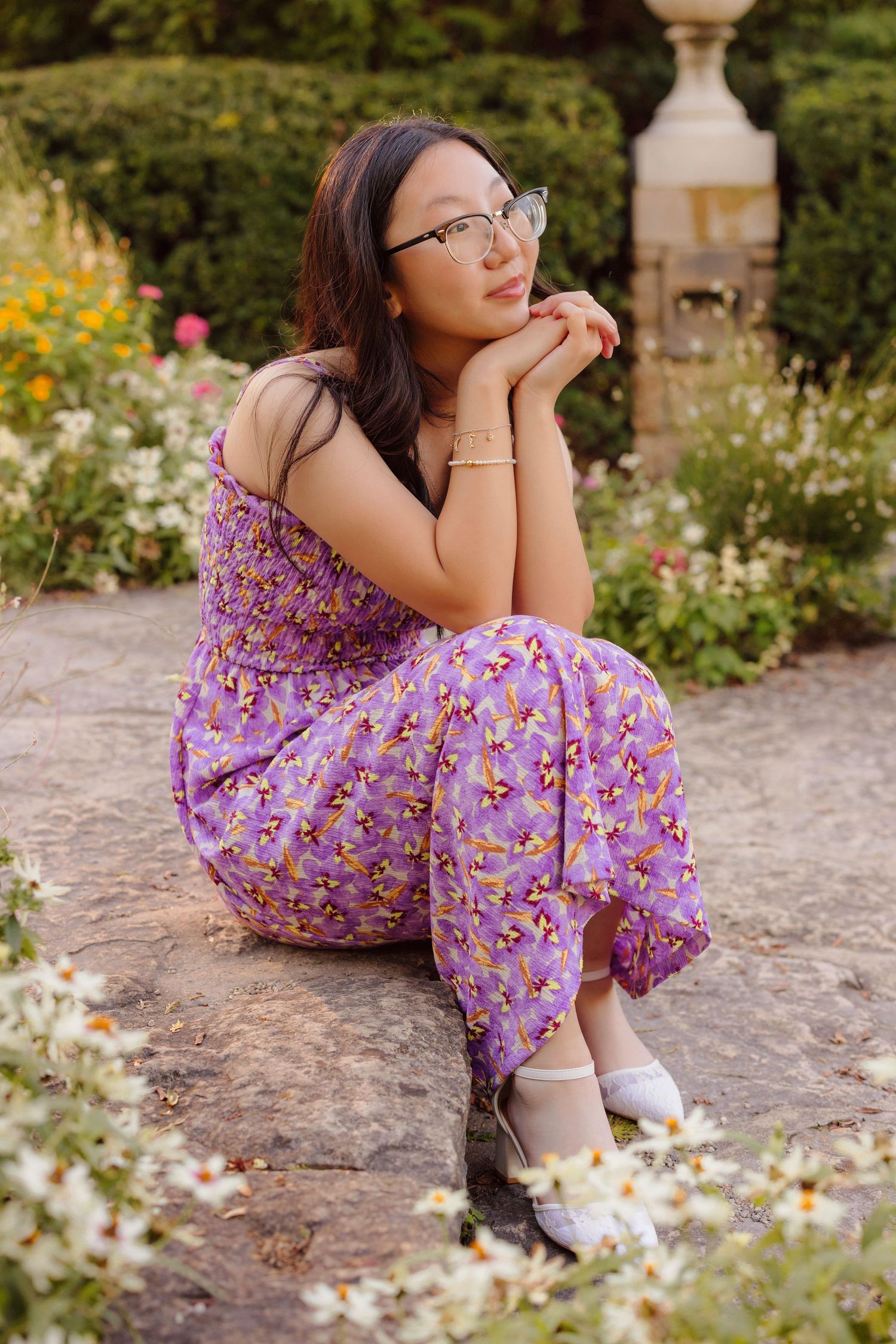 Person in purple floral dress sitting on stone bench surrounded by garden flowers.