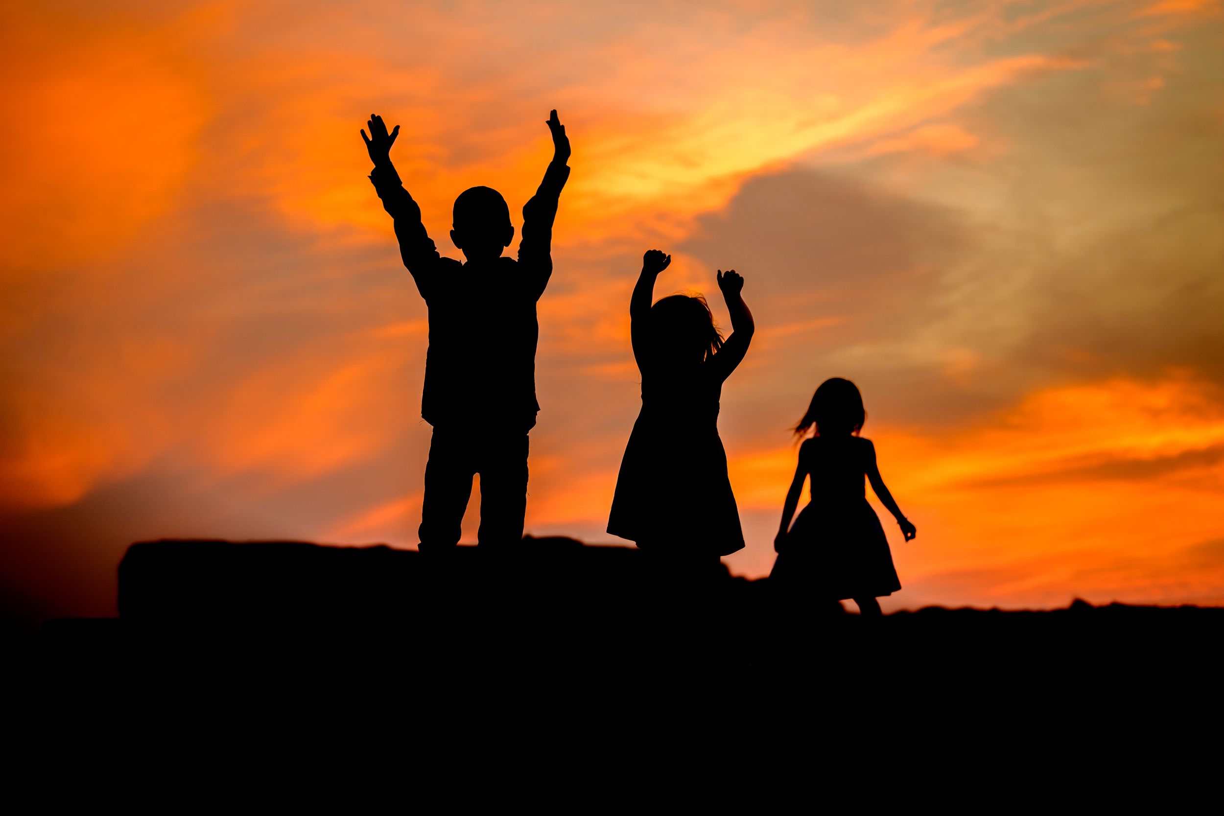 Silhouettes of three children with raised arms standing on a hill against a vibrant orange sunset sky.