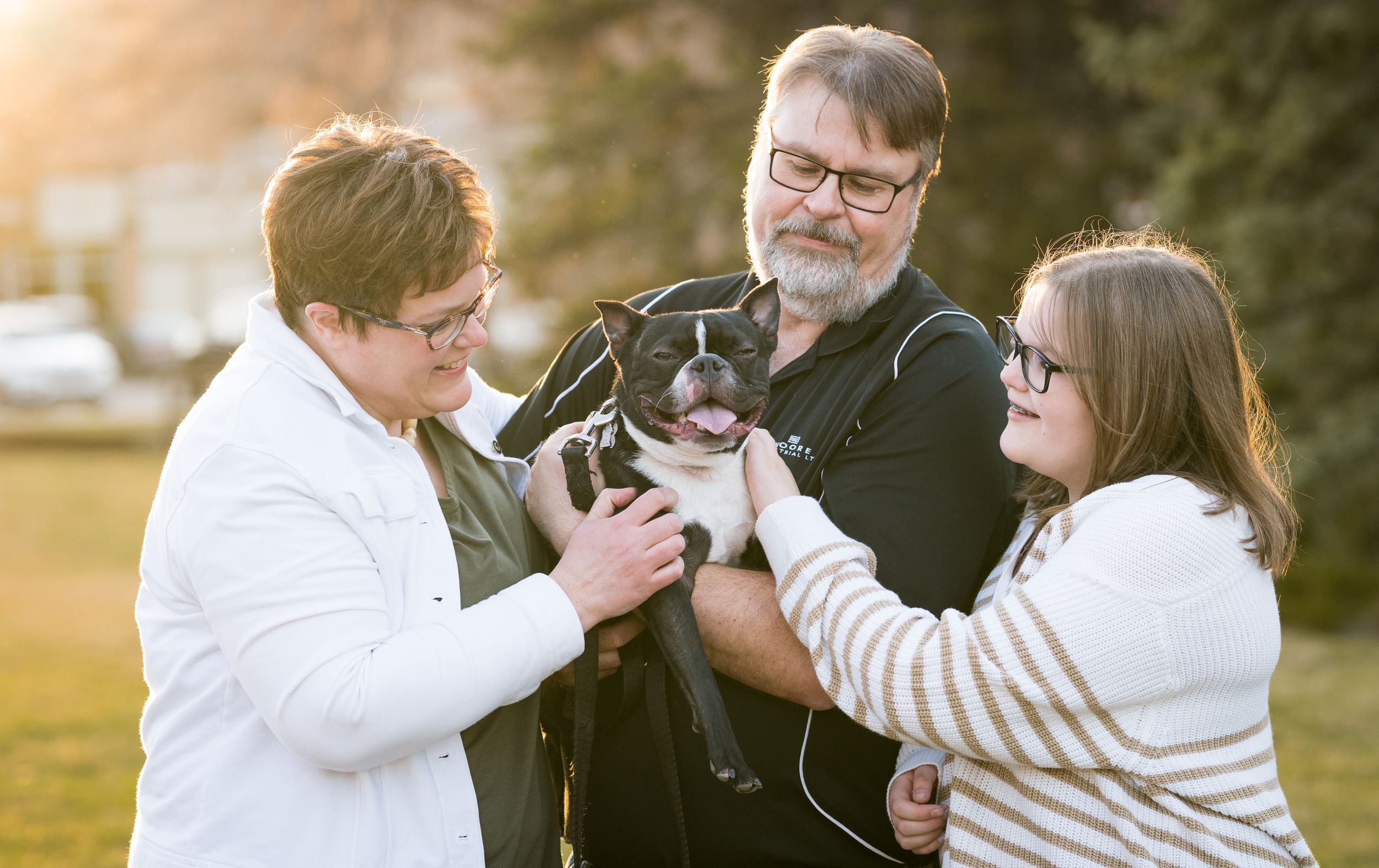 Three people playing with a small dog outdoors during golden hour.