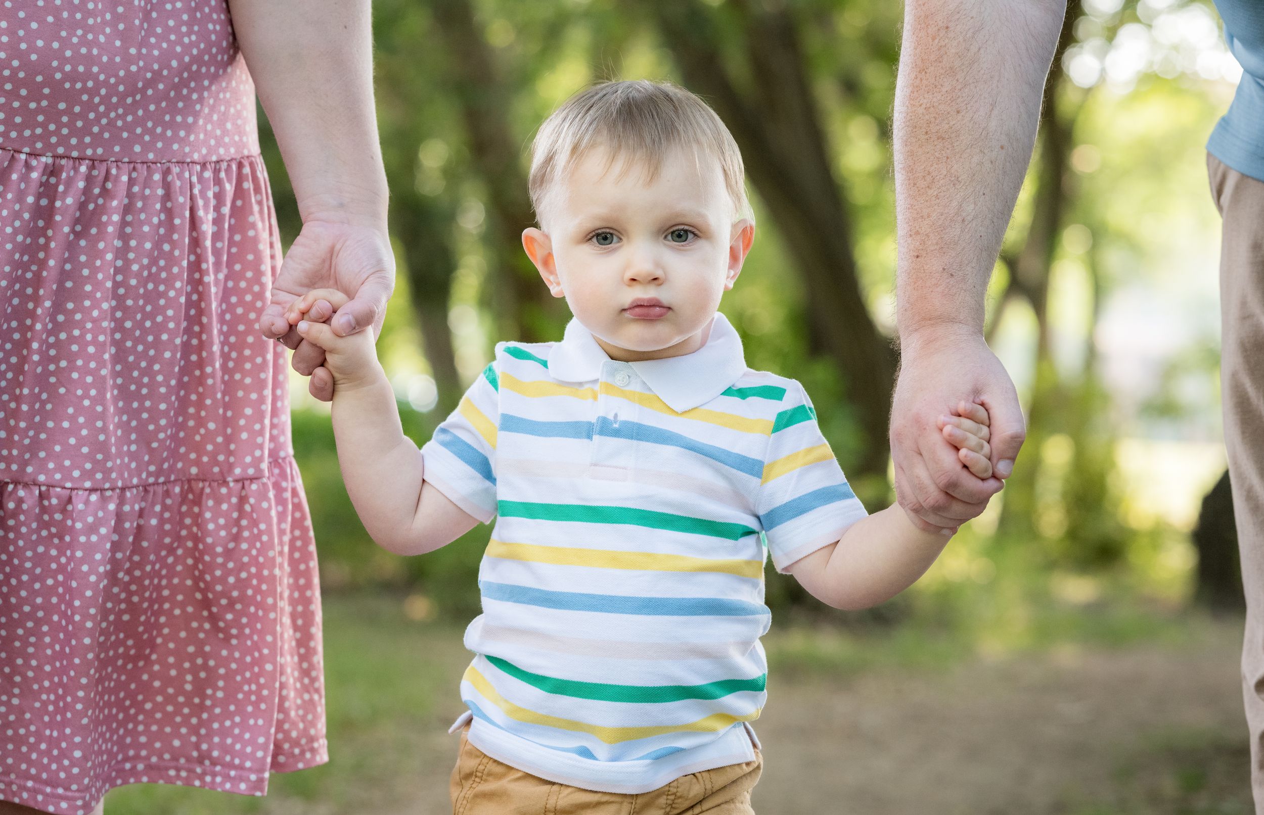 A small child in a striped shirt holds hands with adults while walking in a sunlit forest.