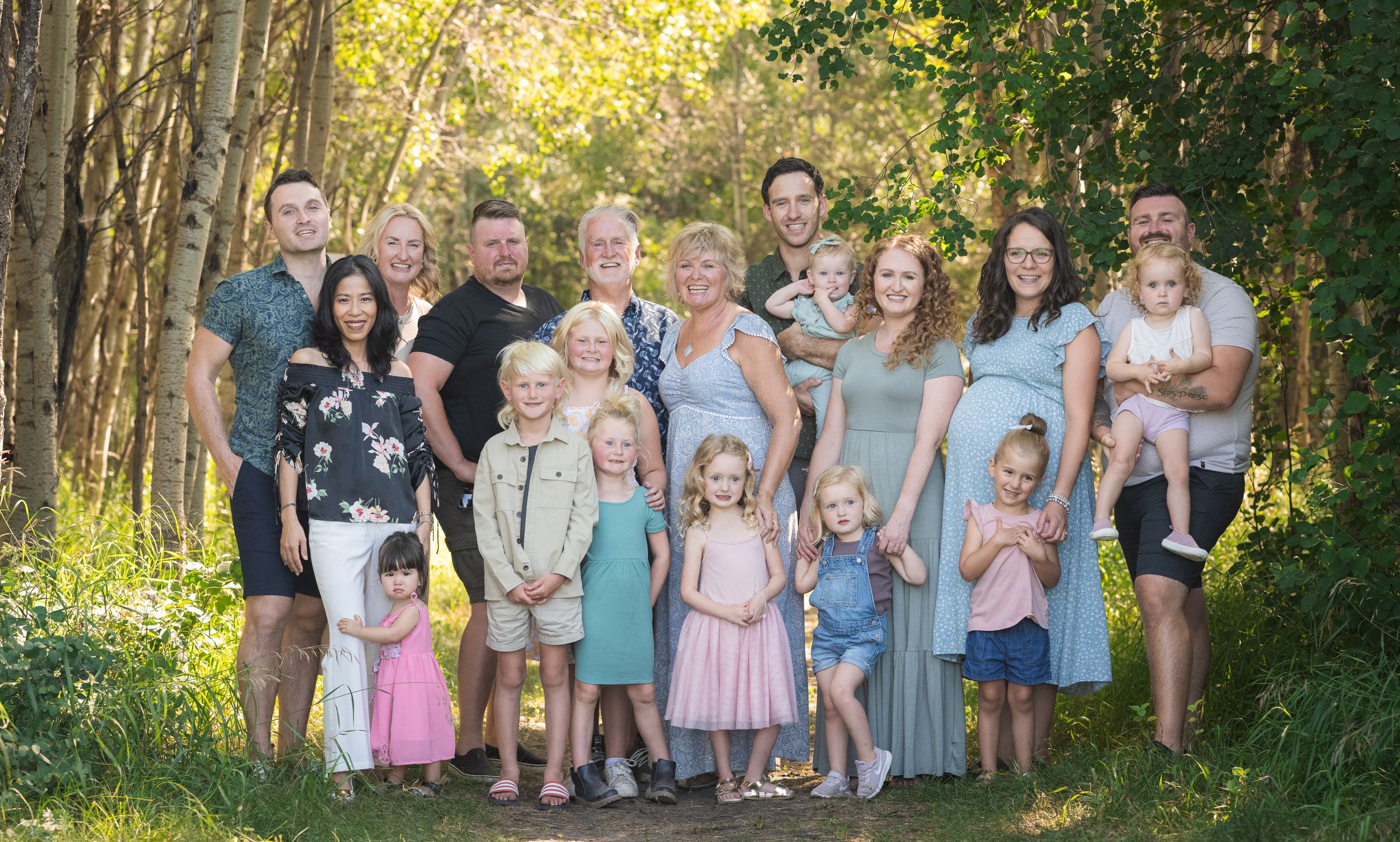 A large extended family gathering poses together outdoors among trees in coordinated outfits.