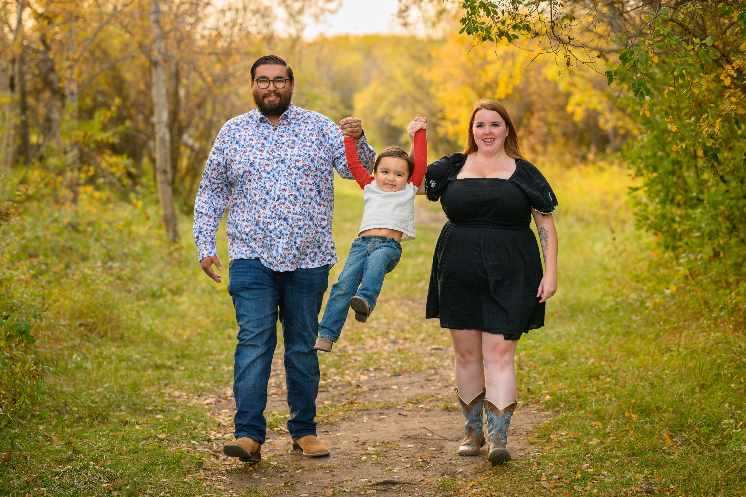 A child swings joyfully between two adults on a dirt path surrounded by golden autumn foliage.