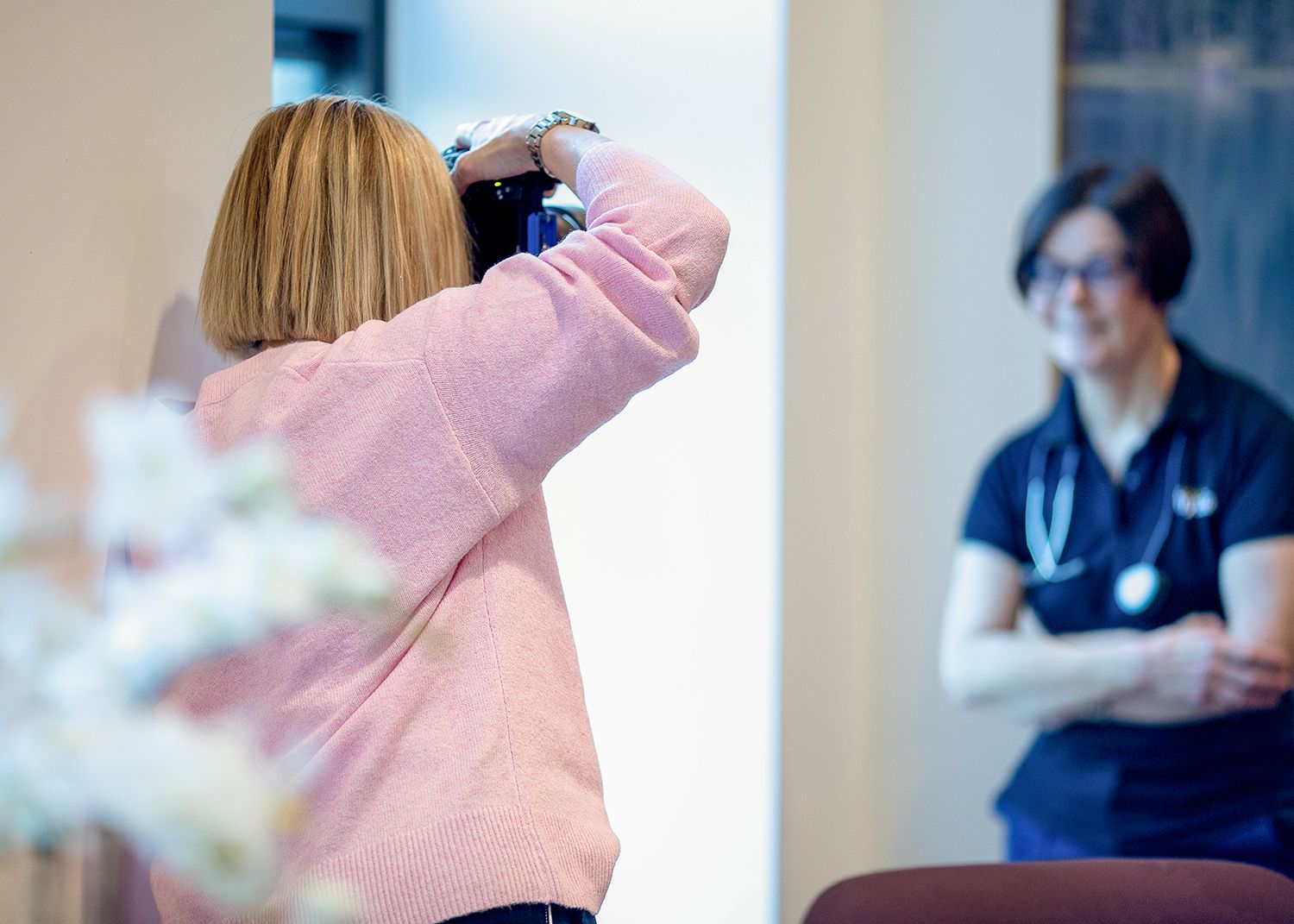A healthcare professional in a pink sweater takes a photo while another medical worker stands in the background.