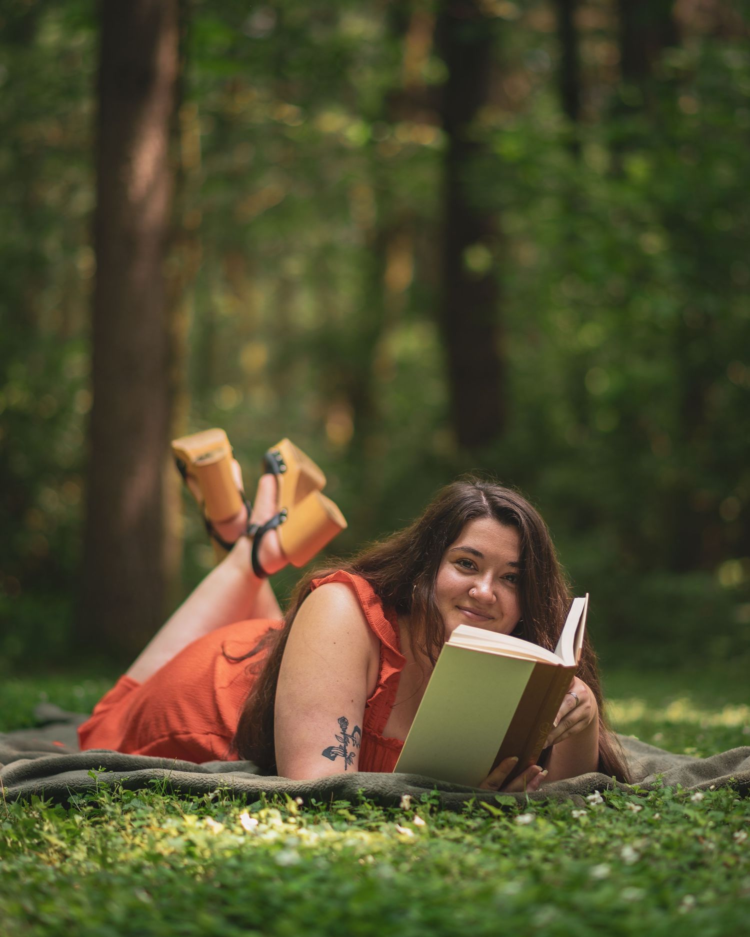 Person in orange dress reads book while relaxing on grass in peaceful forest setting.