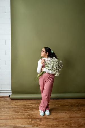 Young girl in pink pants holding white flowers standing against olive green wall.