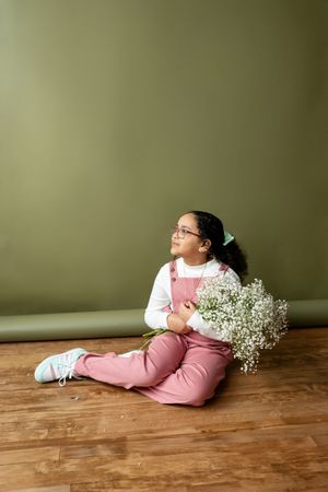 Girl in pink overalls sitting on wooden floor holding white flowers against green wall.