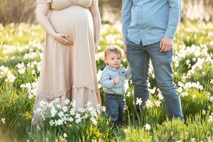 Pregnant woman in pink dress with toddler and partner standing in a field of white daffodils.