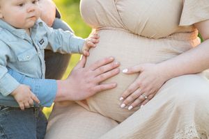 Close-up of hands touching pregnant woman's belly outdoors with toddler nearby.