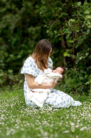 Mother in floral dress sitting in daisy field cradling her newborn baby outdoors.