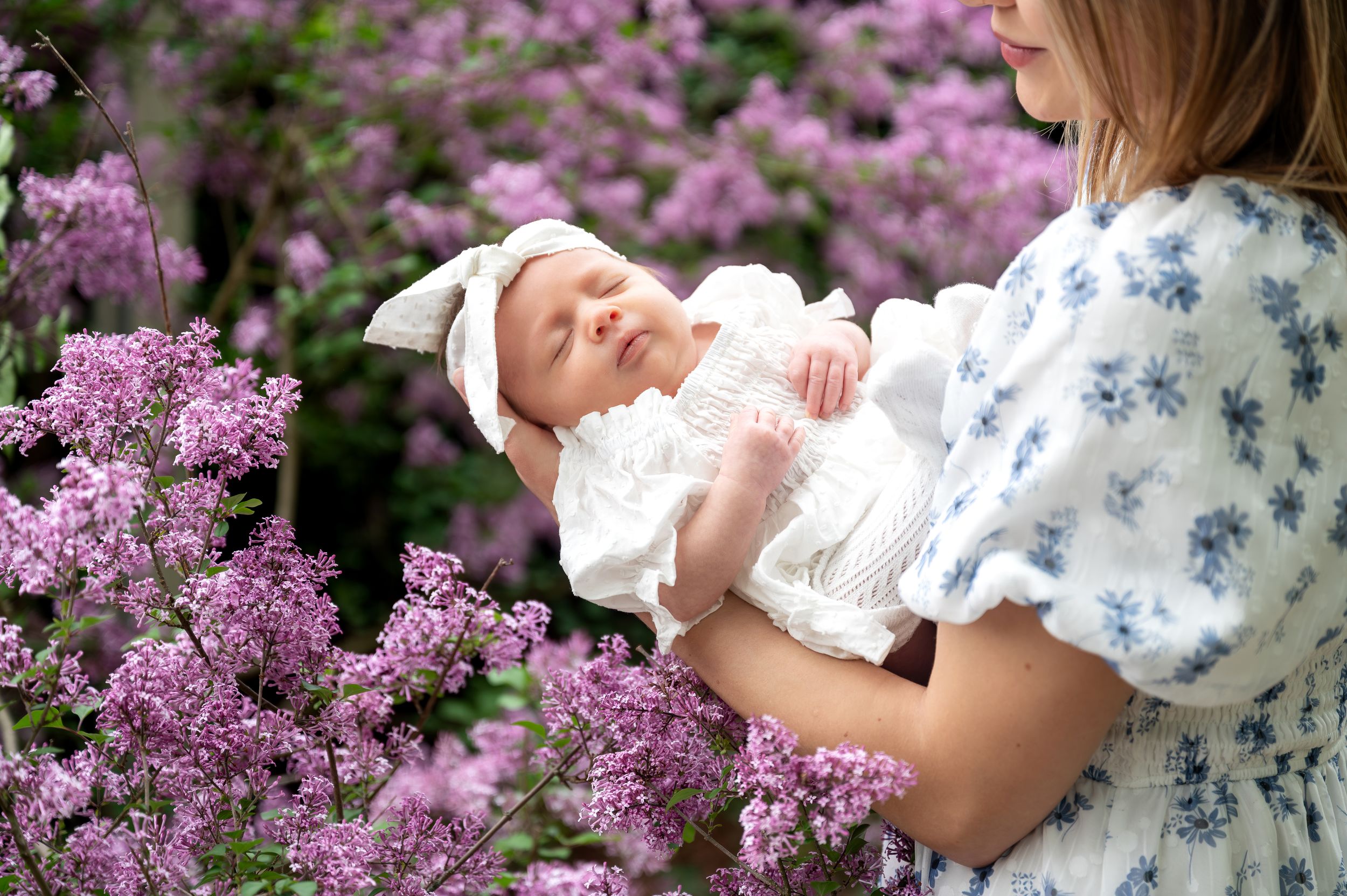Mother holding sleeping newborn baby in white dress among purple lilac flowers outdoors.