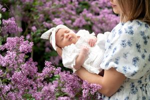 Mother holding sleeping newborn baby in white dress among purple lilac flowers outdoors.