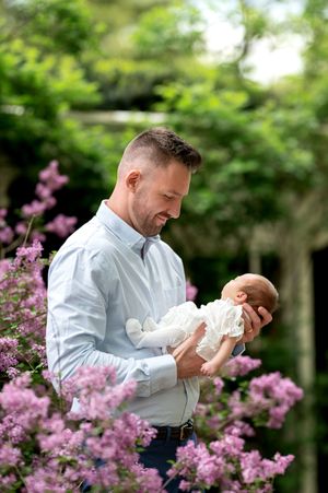 Father tenderly holding newborn baby surrounded by pink flowering bushes in garden.