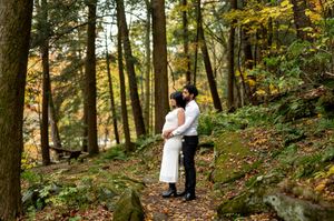 Couple embraces tenderly on a leaf-covered forest path among tall trees with golden autumn leaves.
