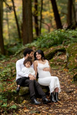 Family of three sits on mossy rocks in an autumn forest, pregnant mother smiling warmly at camera.