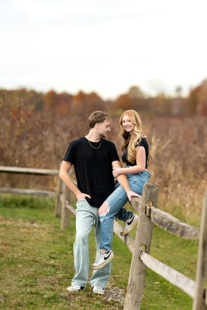 Young couple laughing together near a wooden fence with colorful fall foliage in the background.