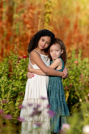 Two young girls embrace warmly in a colorful autumn flower garden.