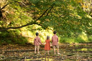 Three barefoot siblings hold hands facing a sunlit autumn forest by a rocky stream.