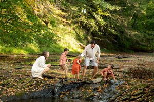A family plays together on rocky terrain beside a stream in a vibrant autumn forest.