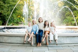 Five young girls smiling and sitting together in front of a large outdoor fountain.