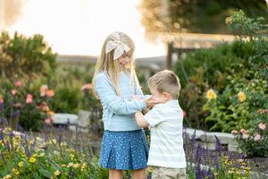 A young girl adjusts her little brother's shirt in a colorful garden with soft golden light.