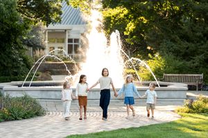 Five children hold hands and walk together near a large fountain in a lush garden.