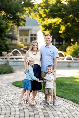 A smiling family of four poses near a fountain, holding a small sign in a park setting.