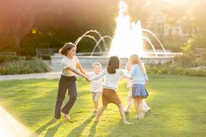 A woman and four children hold hands in a circle on a sunny lawn near a fountain.