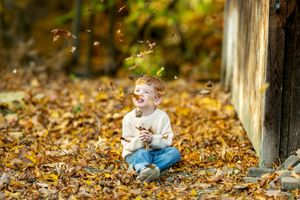 Joyful red-haired toddler laughing while autumn leaves fall around him in golden forest setting.