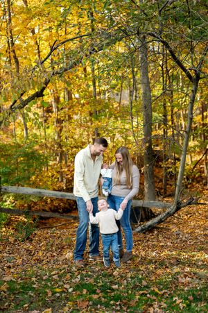 Young family of four holding hands among colorful fallen leaves in golden autumn forest setting.