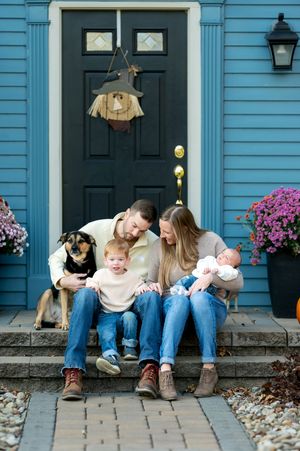 Family with toddler, newborn, and dog sitting on front porch steps of blue house with fall decorations.