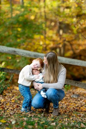 Mother crouching in autumn leaves with toddler son leaning in to kiss newborn baby sibling.