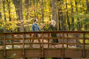 Happy couple walking hand-in-hand across wooden bridge surrounded by vibrant yellow autumn foliage.