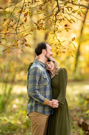 Romantic couple embracing under golden autumn branches, woman in green dress resting head on mans chest.