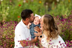 Parents kissing their smiling baby on the cheeks while sitting in a colorful flower field.