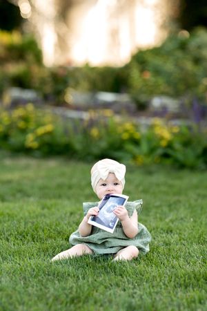 Baby girl in green dress sits on grass holding an ultrasound photo.