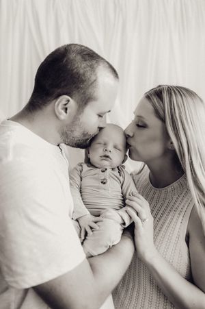 New parents kissing their newborn baby on the cheeks in a tender black and white family portrait.