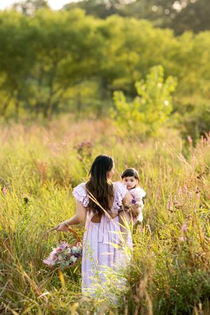 Mother in pink dress carrying her baby and flower basket while walking through a sunlit wildflower meadow.