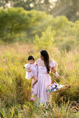Mother and baby daughter in matching pink dresses walking through a golden sunlit meadow with a flower basket.