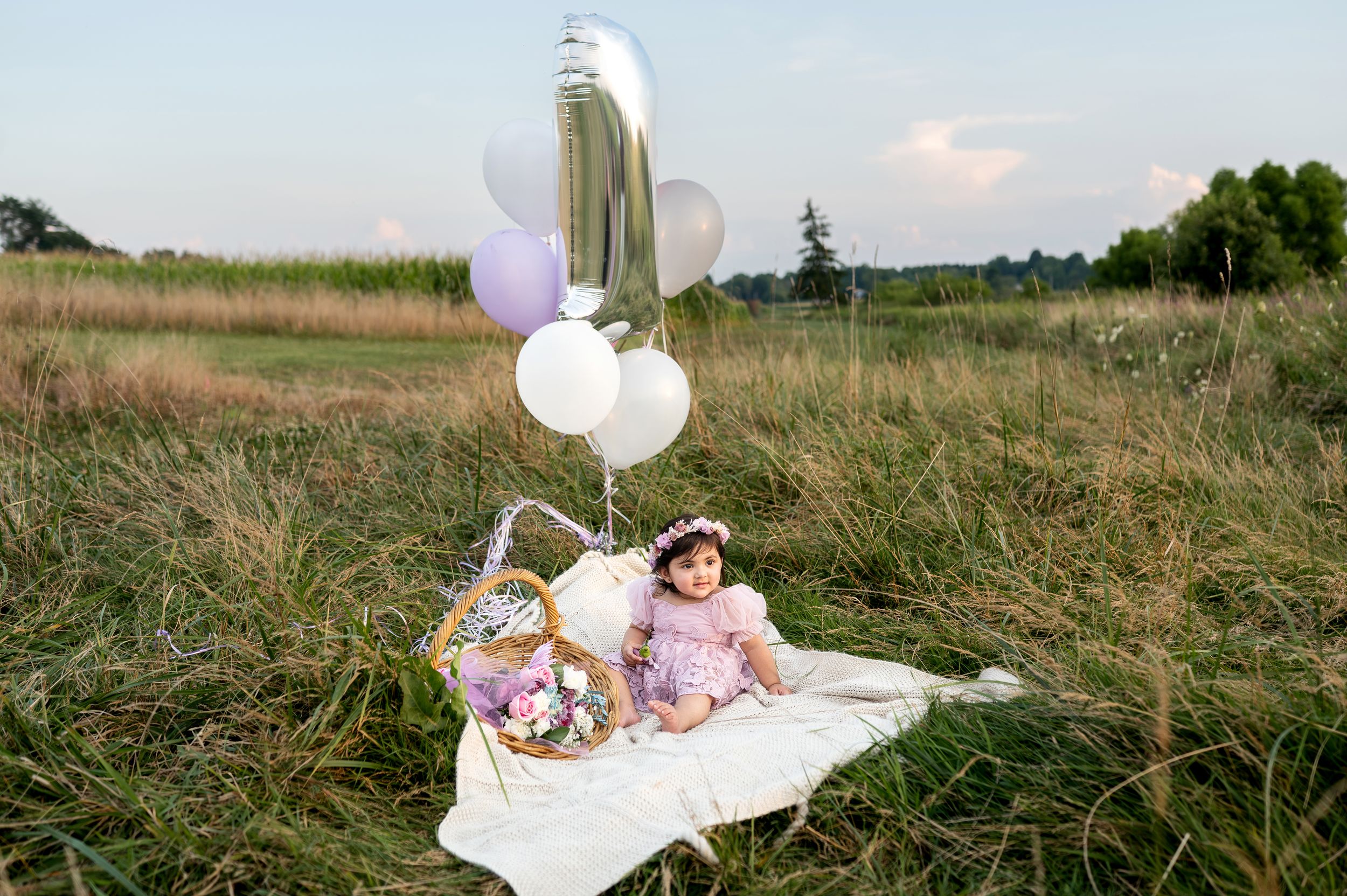 Baby girl in lavender dress sitting on a blanket in a field surrounded by colorful balloons and a flower basket.
