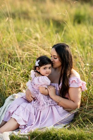 Mother in pink dress kissing her baby girl in a lavender dress while sitting in a golden grass field.
