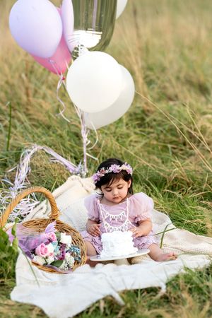 Baby girl in pink dress with floral crown sits on blanket with white cake and balloons outdoors.
