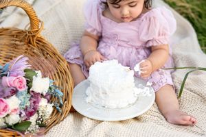 Close-up of baby girl in lavender dress smashing white birthday cake beside flower basket.