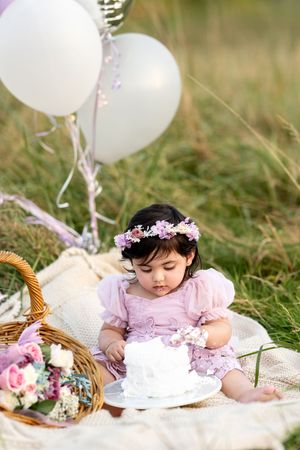 Adorable baby girl in pink dress with flower crown explores white cake surrounded by balloons.