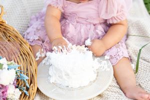 Baby girl in purple dress smashing white birthday cake on plate during outdoor cake smash session.