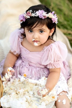 Baby girl with floral crown and frosting on face looks up after smashing white birthday cake.