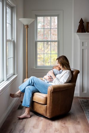 Mother sitting in leather chair holding newborn baby by sunny window at home.