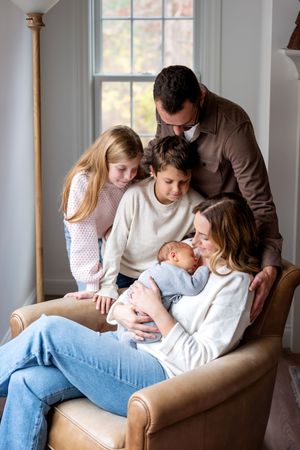 Family of five gathered around mother holding newborn baby in a cozy armchair.